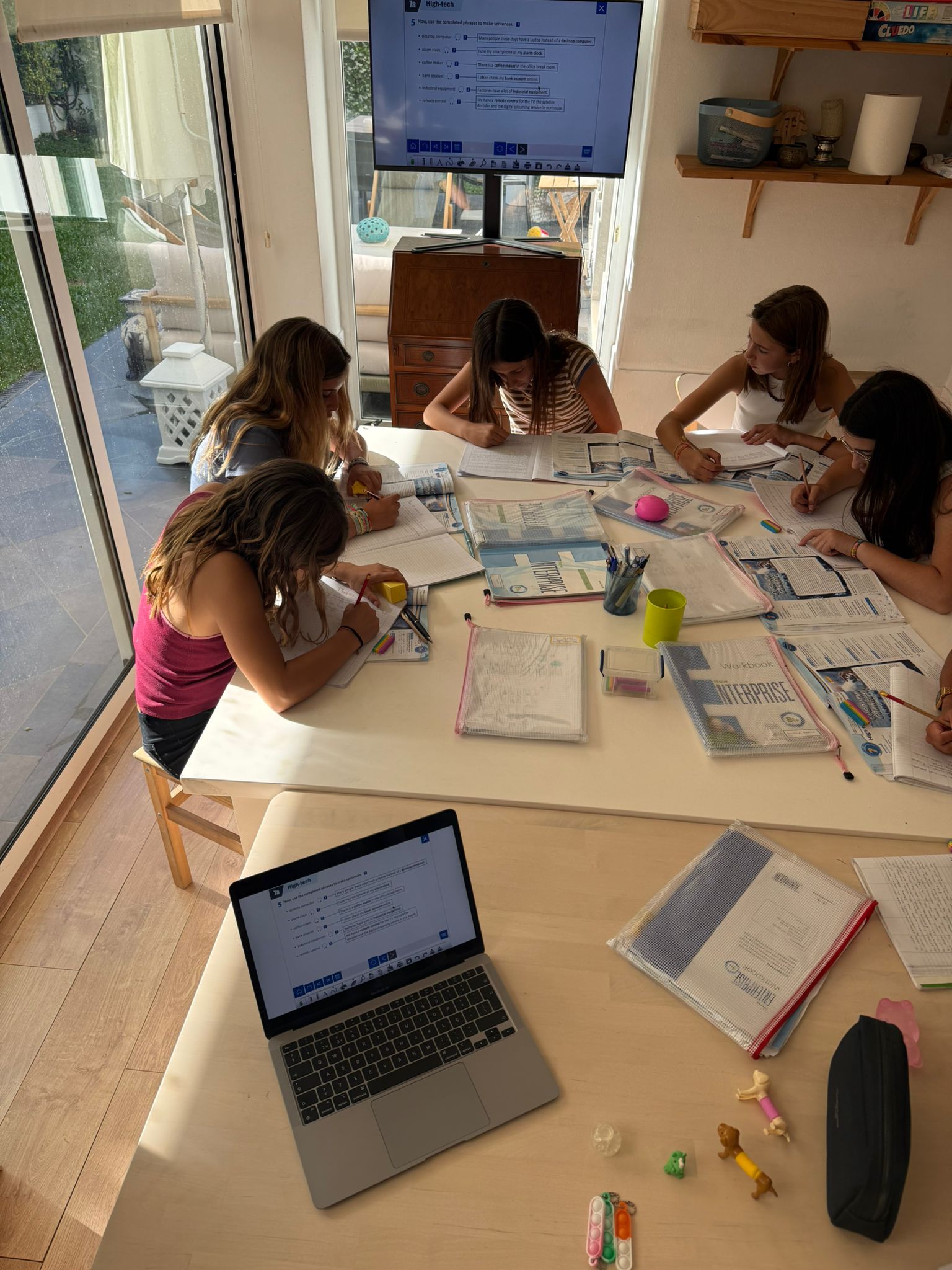Cinco alunas a estudar em torno de uma mesa com manuais, um portátil e um ecrã de TV grande a exibir exercícios